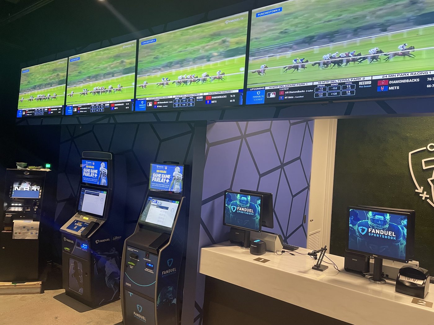 A retail sportsbook counter. A bank of screens above shows live horse racing from several tracks alongside odds and upcoming games; self-service betting kiosks stand below them.