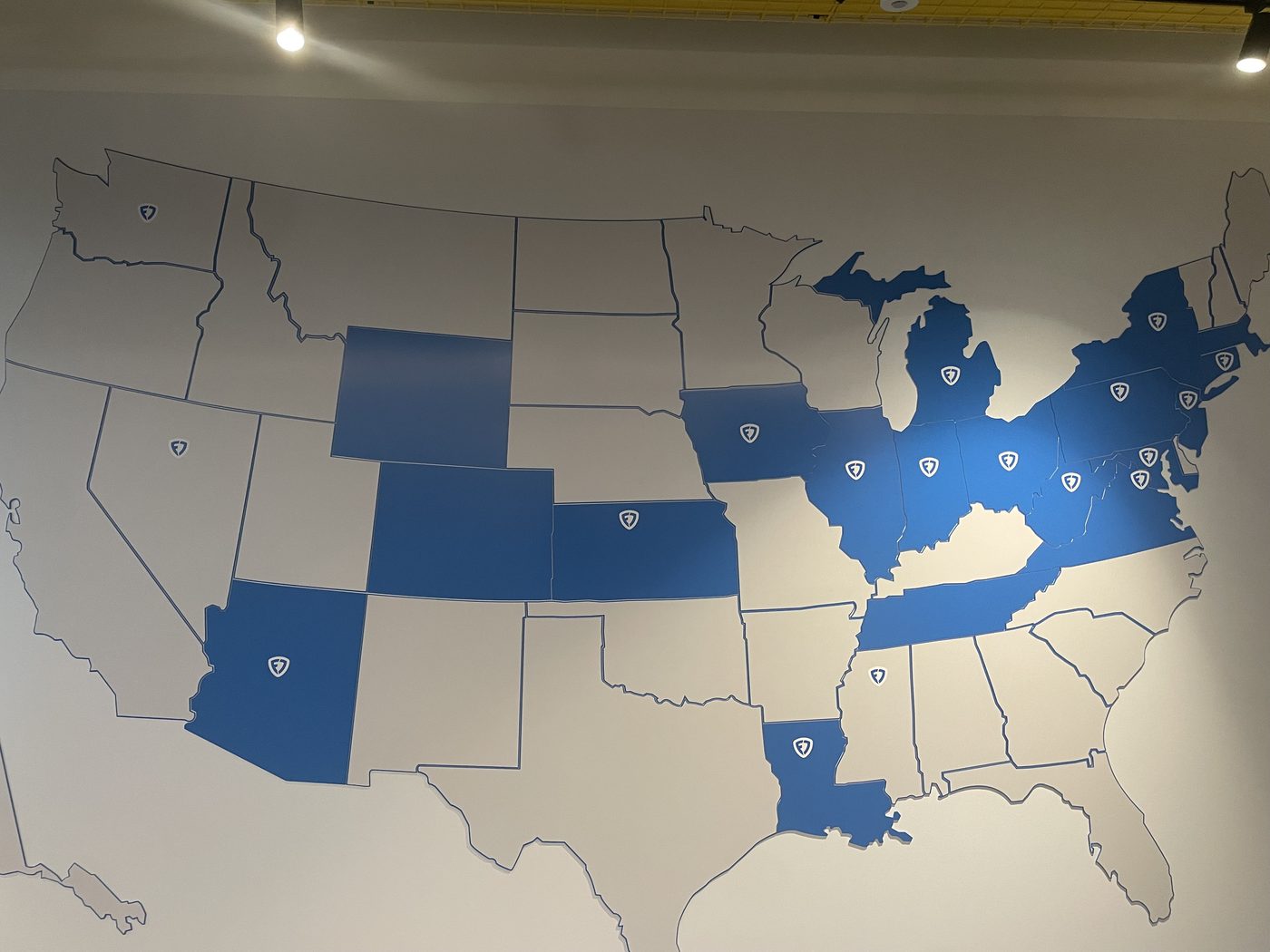 A wall-mounted map of the United States. Roughly twenty states are filled blue and carry a shield; the remainder are blank.