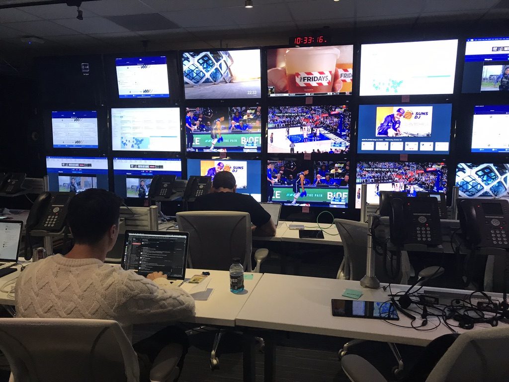 Two people at desks with laptops, desk phones and headsets, facing a wall of monitors showing live NBA games, broadcast feeds and product pages.